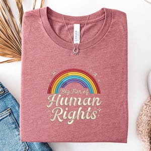 May include: A dusty rose t-shirt with a rainbow graphic and the text "Big Fan of Human Rights." A silver necklace with a sunburst pendant rests on the shirt. The shirt is folded neatly.