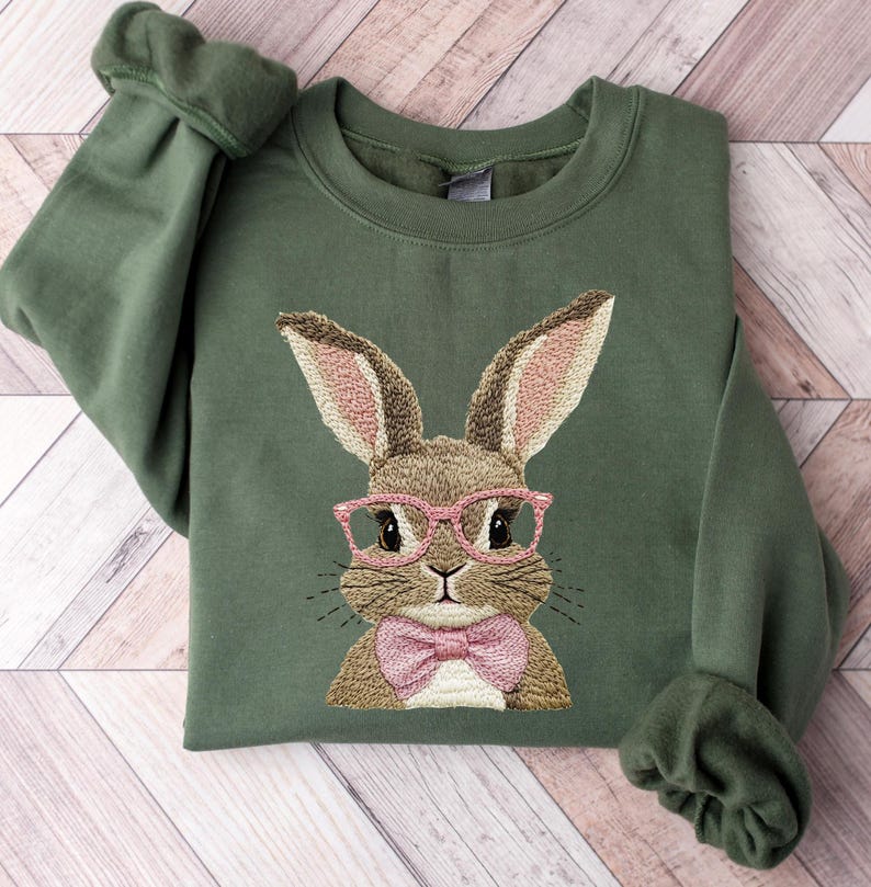 K&ouml;nnte beinhalten: Ein olivgr&uuml;nes Sweatshirt mit einem gestickten Hasen, der eine rosa Brille und eine rosa Fliege tr&auml;gt. Der Hase hat braunes Fell und rosa gef&uuml;tterte Ohren. Das Sweatshirt hat lange &Auml;rmel mit gerippten B&uuml;ndchen und einen Rundhalsausschnitt.