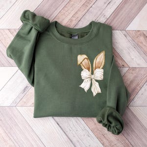 Puede incluir: Una sudadera verde con un dise&ntilde;o de oreja de conejo marr&oacute;n y un lazo blanco.