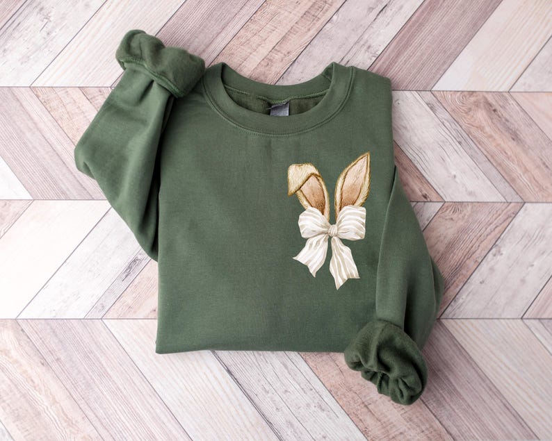 Puede incluir: Una sudadera verde con un dise&ntilde;o de oreja de conejo marr&oacute;n y un lazo blanco.