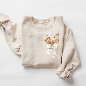 Puede incluir: Una sudadera beige con un dise&ntilde;o de orejas de conejo marrones y blancas en la parte delantera.