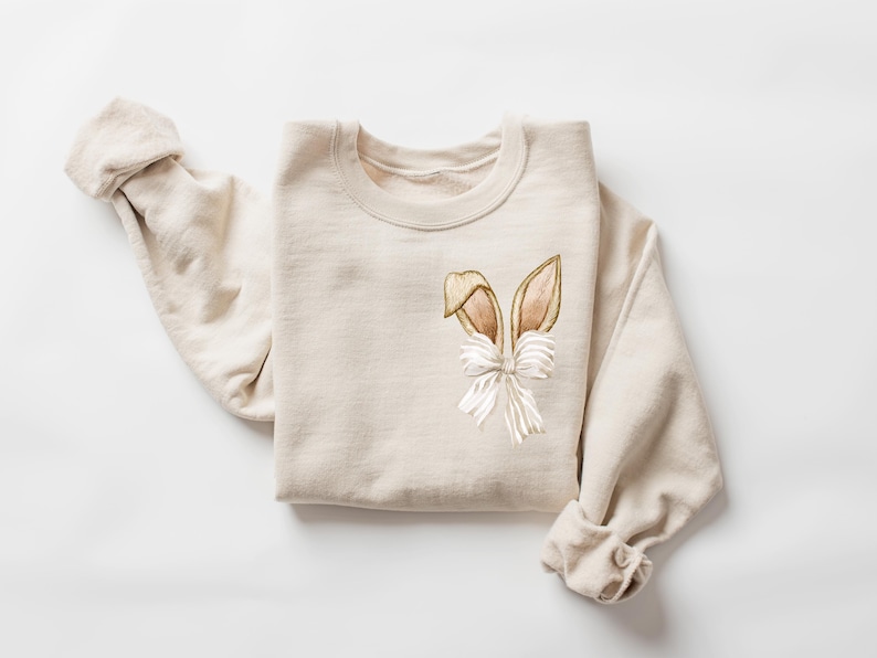 Puede incluir: Una sudadera beige con un dise&ntilde;o de orejas de conejo marrones y blancas en la parte delantera.
