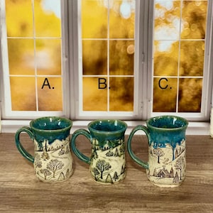 May include: Three ceramic mugs labeled A, B, and C, each featuring a cream-colored base with a hand-drawn black ink landscape of trees, hills, and small cabins, topped with a glossy blue-green glaze that drips down the upper rim.