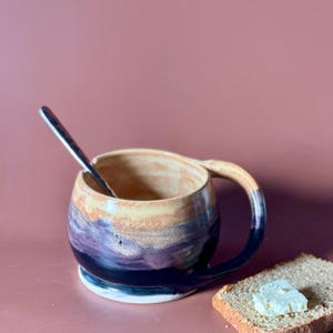 May include: A handmade ceramic mug with a unique color gradient, transitioning from tan to purple and deep blue. The mug has a rounded shape, a sturdy handle, and a silver spoon inside. A slice of bread with butter is next to the mug.