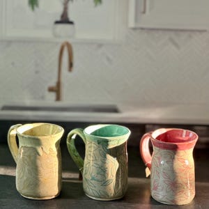 May include: Three handmade ceramic mugs with floral patterns. The mugs have different colors: yellow, green, and red. Each mug has a handle and a unique design. The mugs are displayed on a dark surface, with a kitchen background.