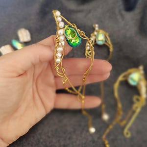 Könnte beinhalten: Ein goldfarbener Draht-Ohrschmuck mit einem grünen Edelstein und weißen Perlen. Das filigrane Design hat eine geschwungene Form mit einem zentralen grünen Juwel und kleinen Perlen, die einen eleganten und ätherischen Look erzeugen. Weitere Ohrschmuck sind im Hintergrund zu sehen.