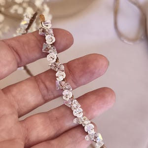 K&ouml;nnte beinhalten: Ein d&uuml;nner, metallischer Haarreif mit einer Reihe abwechselnder, klarer, facettierter Kristallperlen und kleiner, wei&szlig;er, blumenf&ouml;rmiger Elemente. Die Dekorationselemente sind mit d&uuml;nnem Metalldraht am Haarreif befestigt, was ein zartes, strukturiertes Aussehen erzeugt.