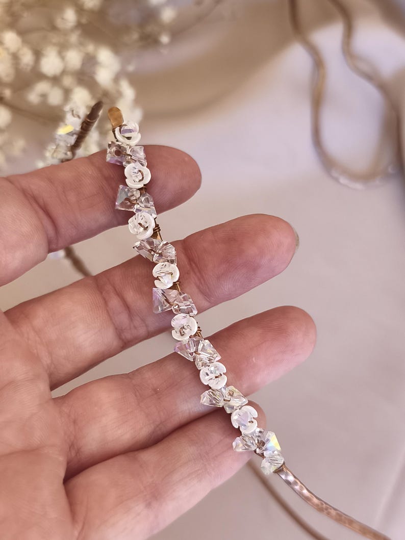 K&ouml;nnte beinhalten: Ein d&uuml;nner, metallischer Haarreif mit einer Reihe abwechselnder, klarer, facettierter Kristallperlen und kleiner, wei&szlig;er, blumenf&ouml;rmiger Elemente. Die Dekorationselemente sind mit d&uuml;nnem Metalldraht am Haarreif befestigt, was ein zartes, strukturiertes Aussehen erzeugt.