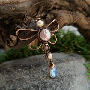 以下が含まれることがあります： ピンクの虹色の石と白い真珠が付いた銅線製のトンボのブローチ。トンボの腹には涙滴型のクリスタルがぶら下がっている。