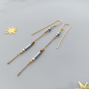 Peut inclure: Une paire de longues boucles d'oreilles pendantes dorées avec des perles géométriques argentées et dorées.