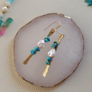 Op de afbeelding: Een paar gouden oorbellen met turquoise en parel accenten. De oorbellen liggen op een witte achtergrond.