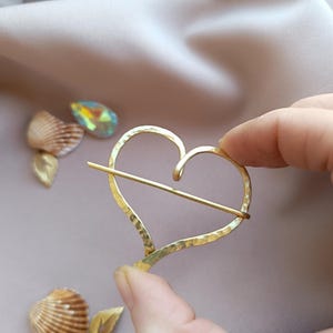 Peut inclure: Épingle à cheveux dorée en forme de cœur avec une texture martelée. La barrette a une barre horizontale au centre. Le fond comprend des coquillages, un cristal et un tissu de couleur neutre.