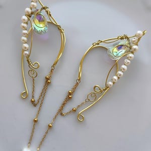 May include: A pair of gold-toned wire ear cuffs featuring a curved frame adorned with a row of small white pearls and a single iridescent teardrop-shaped crystal. Each cuff includes delicate gold chains with small beads and pearl accents.