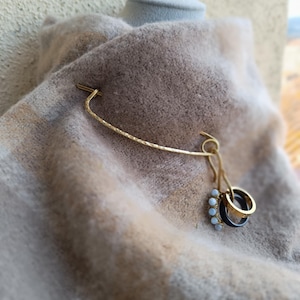 Peut inclure: Épingle à châle dorée avec une texture martelée, tenant une écharpe beige et marron. L'épingle comporte une barre incurvée et un groupe de petits anneaux, dont certains avec des accents bleu clair. L'écharpe est drapée sur un mannequin.