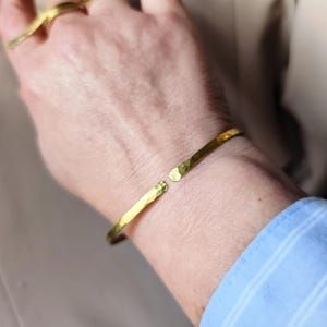 Peut inclure: Un bracelet manchette doré martelé avec un design ouvert. Le bracelet a une surface texturée et un style simple et élégant. Une bague dorée assortie est visible en arrière-plan. La manchette est un accessoire élégant.
