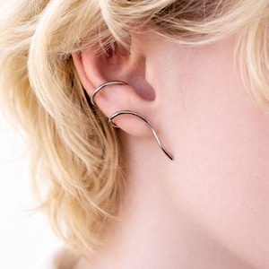 Può includere: Un ear cuff argentato e un ear wrap curvo sono indossati sull'orecchio. L'ear cuff è una semplice fascia, mentre l'ear wrap si curva verso il basso lungo l'orecchio. La persona ha i capelli biondi.