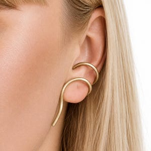 Peut inclure: Une boucle d'oreille en métal doré au fini mat, composée d'un fil incurvé qui entoure le cartilage supérieur et se prolonge vers le bas le long du bord extérieur de l'oreille, présentant une forme métallique continue, fluide et épurée.