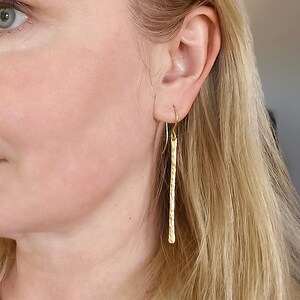 Peut inclure: Gros plan d'une femme portant des boucles d'oreilles en forme de barres en métal doré martelé. Les boucles d'oreilles pendent verticalement et ont une surface texturée. Un collier en or assorti est également visible.