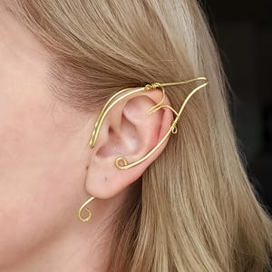 Può includere: Un ear cuff in filo metallico color oro che avvolge il bordo esterno dell'orecchio. Il gioiello presenta una forma appuntita e allungata che si estende verso l'alto e verso l'esterno, con un piccolo dettaglio a spirale sul lobo inferiore.