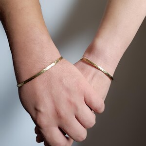 Peut inclure: Deux bracelets en métal doré avec une finition martelée et texturée. Chaque bracelet est un anneau circulaire fin porté autour du poignet. Le métal présente une surface réfléchissante et polie qui capte la lumière, soulignant le motif irrégulier.