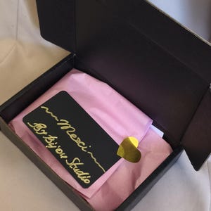 K&ouml;nnte beinhalten: Eine schwarze Geschenkbox aus Pappe mit hellrosa Seidenpapier. Eine schwarze rechteckige Karte mit goldenem Schreibschrifttext Merci By bijou Studio liegt obenauf, befestigt mit einem kleinen, gl&auml;nzenden goldenen herzf&ouml;rmigen Aufkleber.