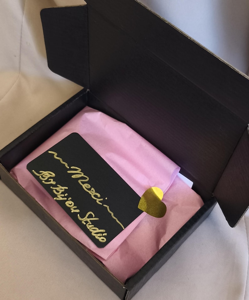 K&ouml;nnte beinhalten: Eine schwarze Geschenkbox aus Pappe mit hellrosa Seidenpapier. Eine schwarze rechteckige Karte mit goldenem Schreibschrifttext Merci By bijou Studio liegt obenauf, befestigt mit einem kleinen, gl&auml;nzenden goldenen herzf&ouml;rmigen Aufkleber.