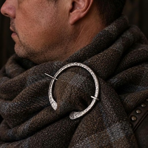 May include: A circular silver-toned brooch with a hammered, textured finish, featuring a thin, straight pin that passes through the centre to secure a thick, brown and grey plaid woven fabric wrap in place.