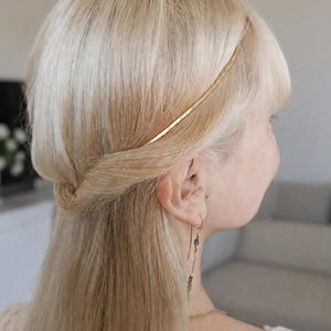 May include: A thin, flat gold-toned metal headband worn across the top of blonde hair. The hair is styled in a half-up, half-down fashion with a subtle twist at the side, complemented by a long, dangling gold-toned earring with small accents.