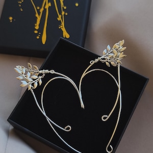 Peut inclure: Des manchons d'oreille argentés avec des accents de feuilles, présentés dans une boîte noire. Les manchons d'oreille ont un design délicat en forme de fil qui épouse l'oreille. La boîte a un couvercle noir avec des éclaboussures de peinture jaune.