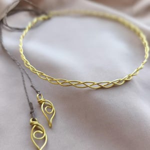 May include: A gold-colored braided headband with two matching dangling charms. The headband and charms are made of thin wire and are displayed on a soft, neutral-toned fabric background. The charms have a teardrop shape with a spiral design.