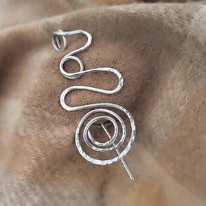 Peut inclure: Une épingle à cheveux en métal argenté avec une texture martelée. Le design se compose d'un fil continu formé en une série de boucles interconnectées et d'une grande spirale à la base, avec une tige droite sortant du centre de la spirale.