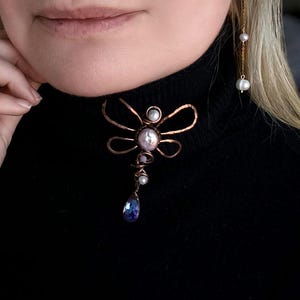 Può includere: Una collana con ciondolo a forma di libellula in filo di rame con una grande perla centrale e un cristallo blu a goccia. La collana è indossata sopra un dolcevita nero. È visibile anche un orecchino a catena d'oro con perle.