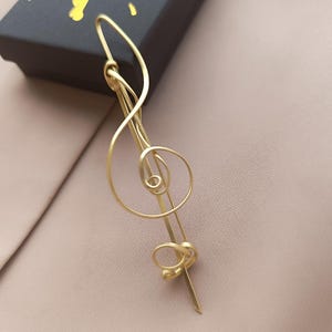 Peut inclure: Une épingle à cheveux dorée en forme de clé de sol. L'épingle présente un design incurvé avec une spirale et un détail de nœud. Elle repose sur une surface rose clair, avec une boîte noire en arrière-plan.
