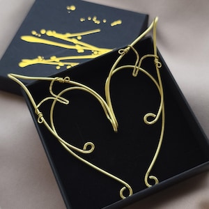 Peut inclure: Une paire d'oreilles d'elfe en fil d'or, présentée dans une boîte noire ouverte. Les manchettes d'oreille présentent un motif en forme de cœur avec des volutes et des courbes complexes. La boîte a un motif abstrait doré sur le couvercle.