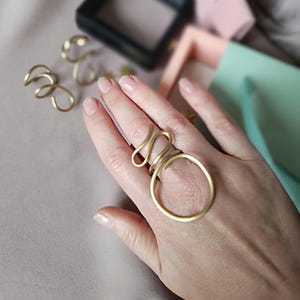 Peut inclure: Un ensemble de bagues dorées, l'une avec un grand motif circulaire et l'autre avec un motif plus complexe et incurvé. Les bagues sont portées sur une main.