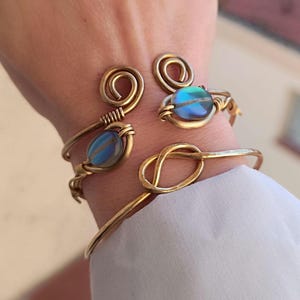 Puede incluir: Tres pulseras de alambre dorado con piedras azules iridiscentes. Las pulseras están apiladas en la muñeca de una persona.