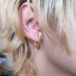 Peut inclure: Boucles d'oreilles créoles dorées à double boucle. Les boucles d'oreilles sont portées à l'oreille et se détachent sur des cheveux blonds clairs. Elles présentent une finition lisse et polie, avec un style simple et élégant.