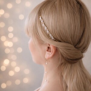 K&ouml;nnte beinhalten: Ein d&uuml;nnes, goldfarbenes Haarband mit kleinen wei&szlig;en Blumenakzenten und klaren Kristallen, kombiniert mit einem goldfarbenen Ohrh&auml;nger, der aus einer d&uuml;nnen Kette und drei kleinen wei&szlig;en, kugelf&ouml;rmigen Perlen an einem Haken besteht.