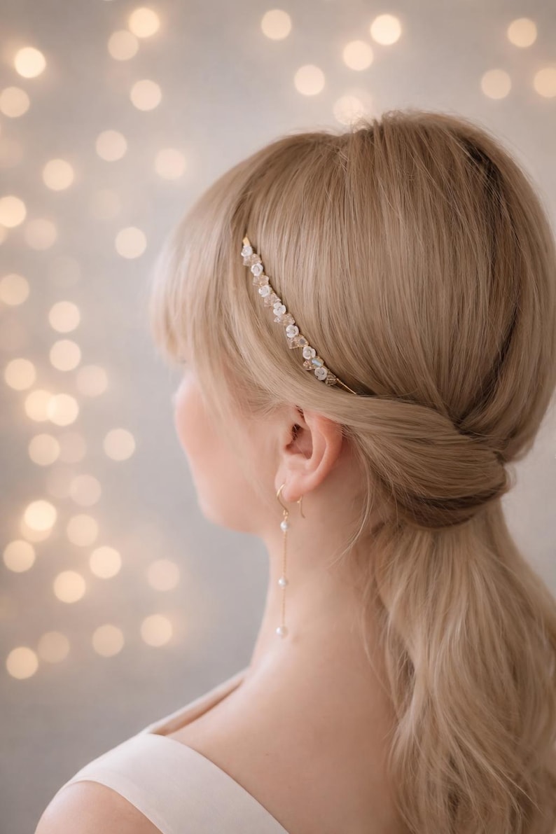K&ouml;nnte beinhalten: Ein d&uuml;nnes, goldfarbenes Haarband mit kleinen wei&szlig;en Blumenakzenten und klaren Kristallen, kombiniert mit einem goldfarbenen Ohrh&auml;nger, der aus einer d&uuml;nnen Kette und drei kleinen wei&szlig;en, kugelf&ouml;rmigen Perlen an einem Haken besteht.