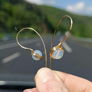 Puede incluir: Un par de pendientes de alambre dorados. Cada pendiente presenta un diseño en forma de lágrima con una pequeña cuenta redonda y opalina. Las cuentas están envueltas con alambre dorado. El fondo está borroso, mostrando una carretera y vegetación.