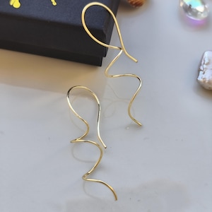 Peut inclure: Une paire de boucles d'oreilles dorées de forme abstraite. Les boucles d'oreilles ont un design incurvé et fluide, avec un crochet en haut pour l'oreille. Les boucles d'oreilles sont présentées sur une surface de couleur claire.