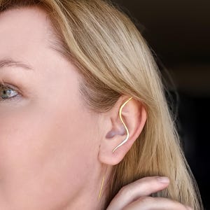 May include: A gold ear cuff and earring set. The ear cuff curves along the upper ear, while a straight earring dangles below. The jewelry is worn on a person with blonde hair and fair skin. The background is blurred.