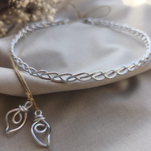 Puede incluir: Un collar tipo gargantilla de alambre plateado con un patrón trenzado. El collar incluye un cordón fino de color tostado con dos dijes de alambre colgantes, cada uno con forma de bucle de gota con un centro en espiral.