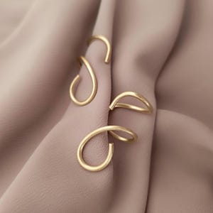 Può includere: Due orecchini a cerchio aperti dorati intrecciati. Gli orecchini sono su uno sfondo di tessuto marrone chiaro.