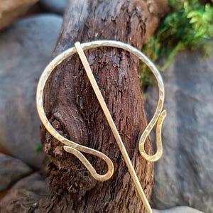 Puede incluir: Un broche de metal dorado con un acabado martillado. El broche tiene forma de círculo con dos bucles en forma de lágrima en la parte inferior. Un alfiler largo y recto está unido al broche.