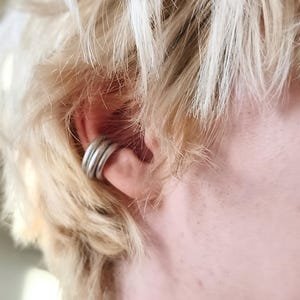 Peut inclure: Un ear cuff argenté composé de trois anneaux au fini lisse et arrondi. Ce bijou est placé sur le cartilage de l'oreille, présentant une structure métallique simple qui s'enroule autour du bord extérieur de l'oreille de manière fixe.