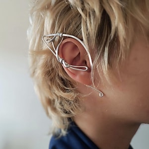 Op de afbeelding: Een zilveren oor manchet, ontworpen om op een elfen oor te lijken, wordt op het oor gedragen. De oor manchet is gemaakt van dun, gebogen metaaldraad, met een gebogen ontwerp dat rond het oor loopt. De oor manchet is een uniek sieraad.