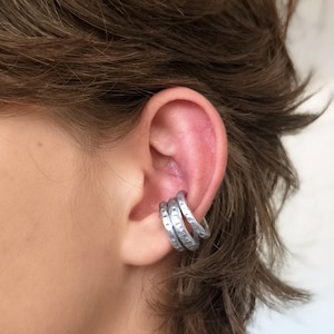 Peut inclure: Un ear cuff argenté composé de trois anneaux parallèles avec une finition martelée et texturée. Ce bijou se place sur la conque de l'oreille, créant un effet superposé grâce à ses trois bandes métalliques distinctes et régulières.