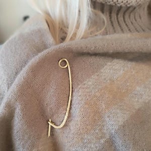 Puede incluir: Un broche de alfiler de seguridad dorado con un diseño curvo. El alfiler está sujeto a un suéter beige. El broche tiene un diseño simple y elegante, adecuado para agregar un toque de estilo a cualquier atuendo.