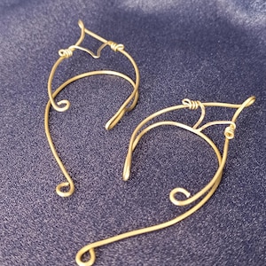 May include: A pair of gold-coloured ear cuffs, crafted from wire and designed to sit around the ear. The jewellery features a curved design with a decorative top and a curled base. Set against a dark blue fabric.